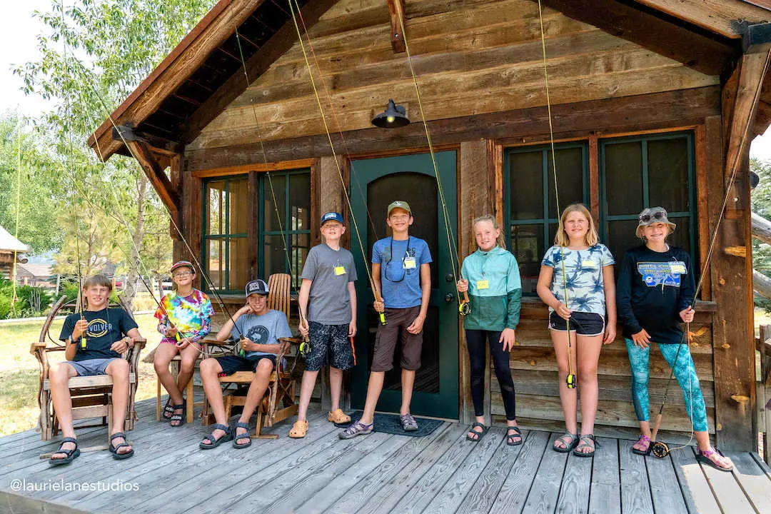 A group of children stand on a wooden porch with fly fishing rods.