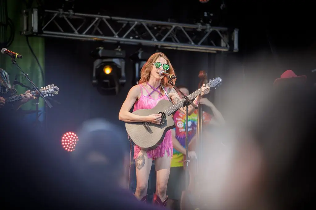 A woman performs on stage with an acoustic guitar while other musicians play behind her.