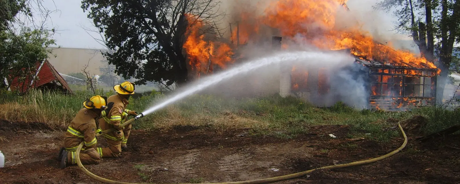 Two firefighters are kneeling on the ground while spraying water at a burning building.