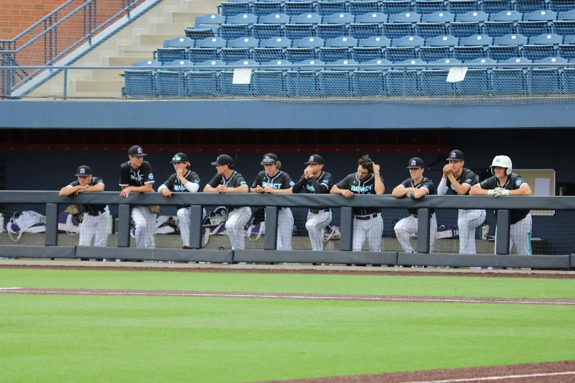 A group of baseball players in uniforms lean against the dugout railing at a baseball field.