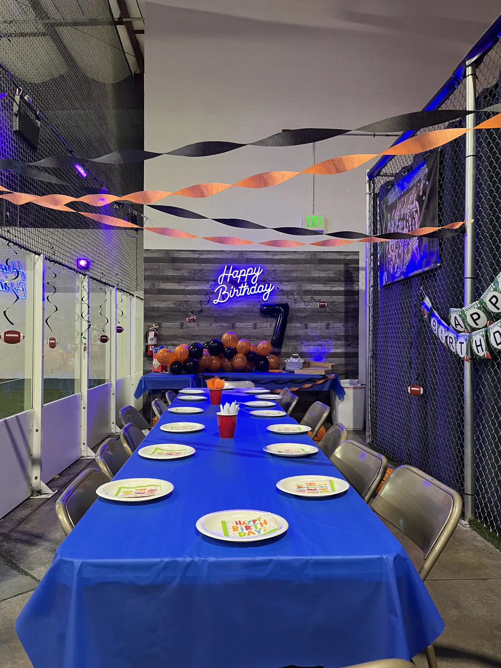 A birthday party table is decorated with a blue tablecloth, plates, and balloons in an indoor sports facility.