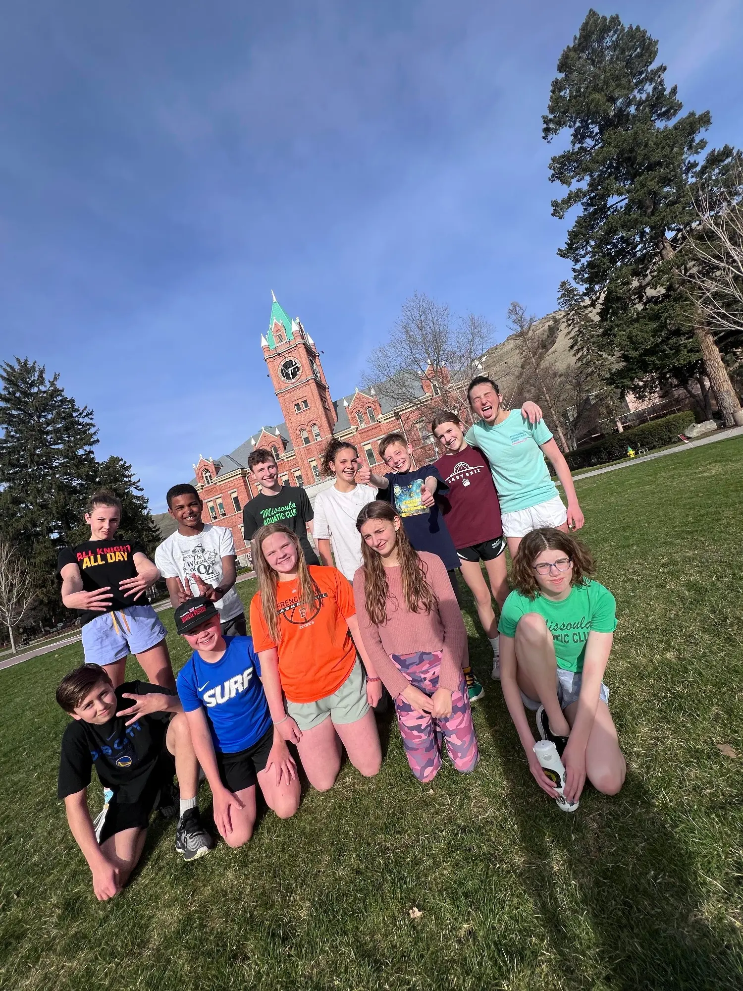 A group of young people poses together on a grassy lawn in front of a historic brick building.