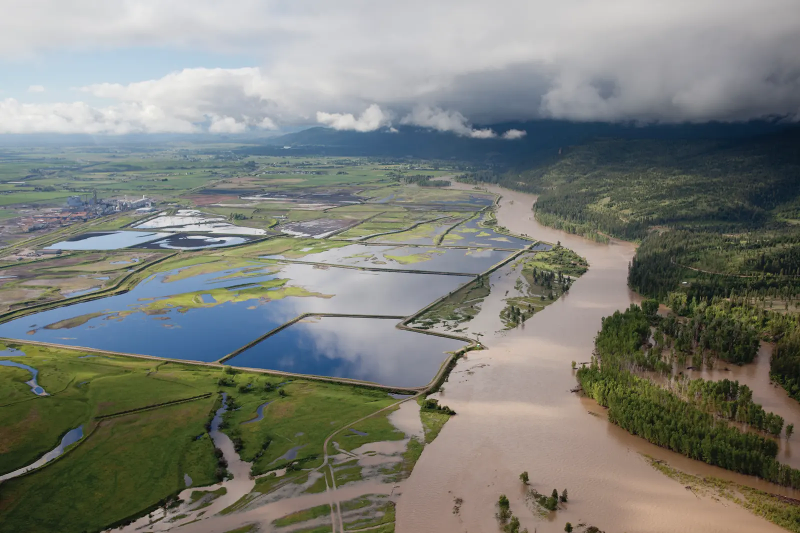 An aerial view shows a large river flowing alongside industrial ponds and fields.