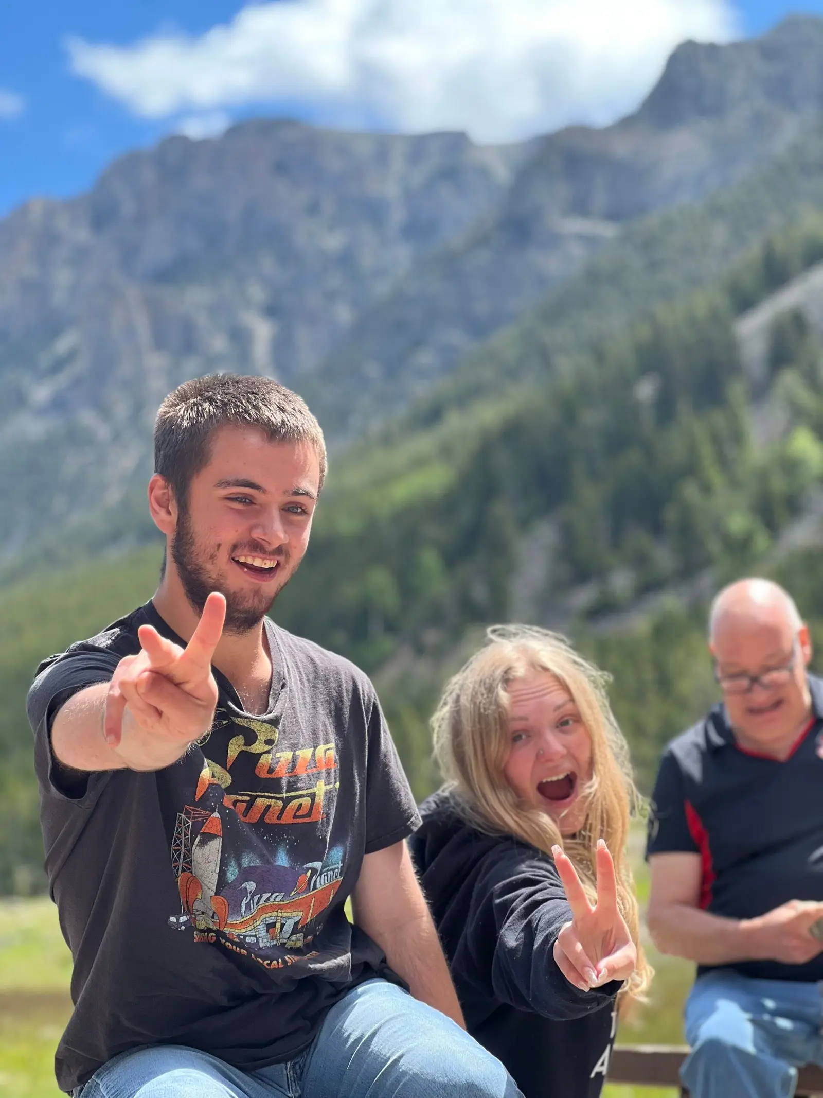 Two people pose with peace signs in front of a mountainous background.