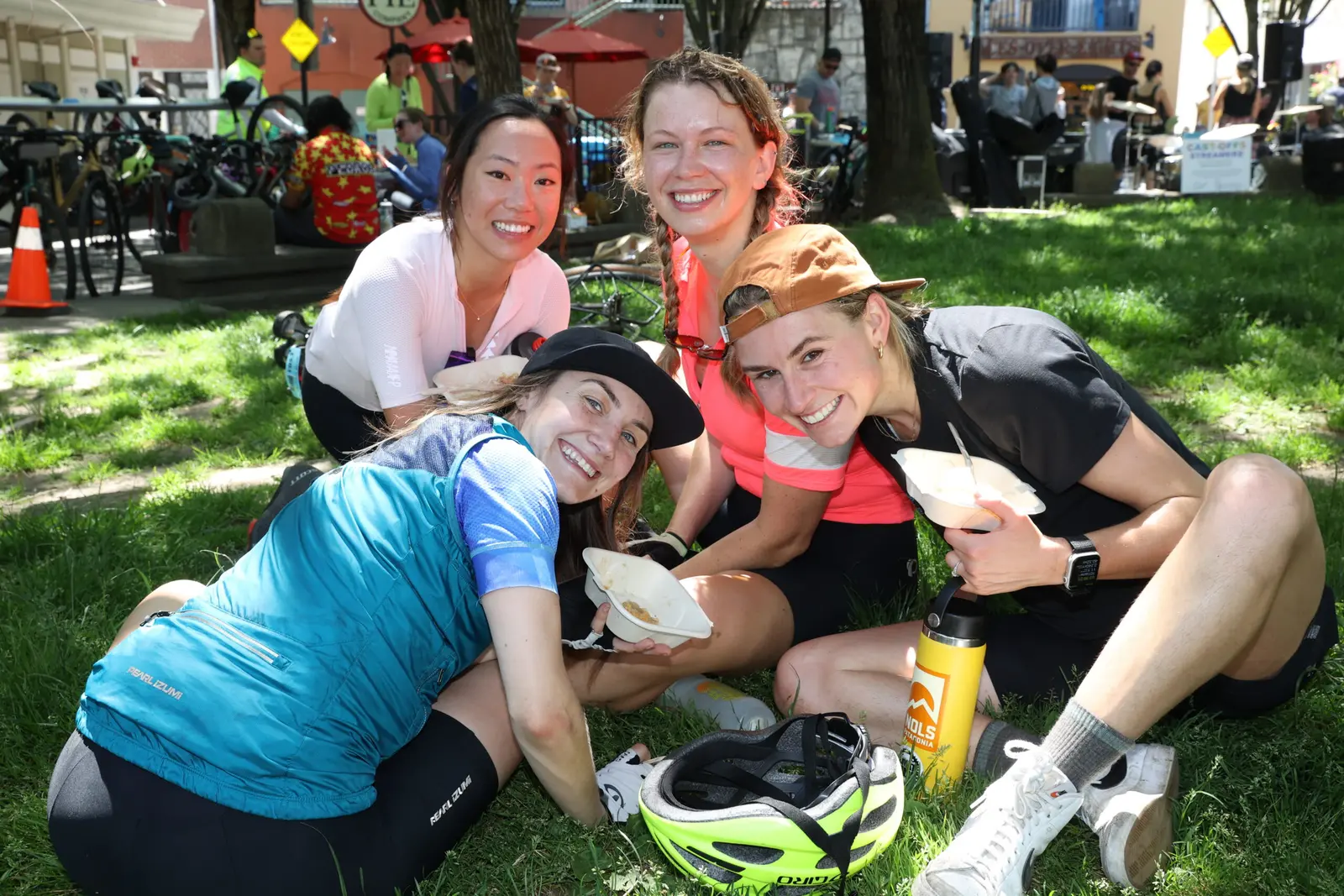 Four young women sit on the grass outdoors and smile while holding food containers.