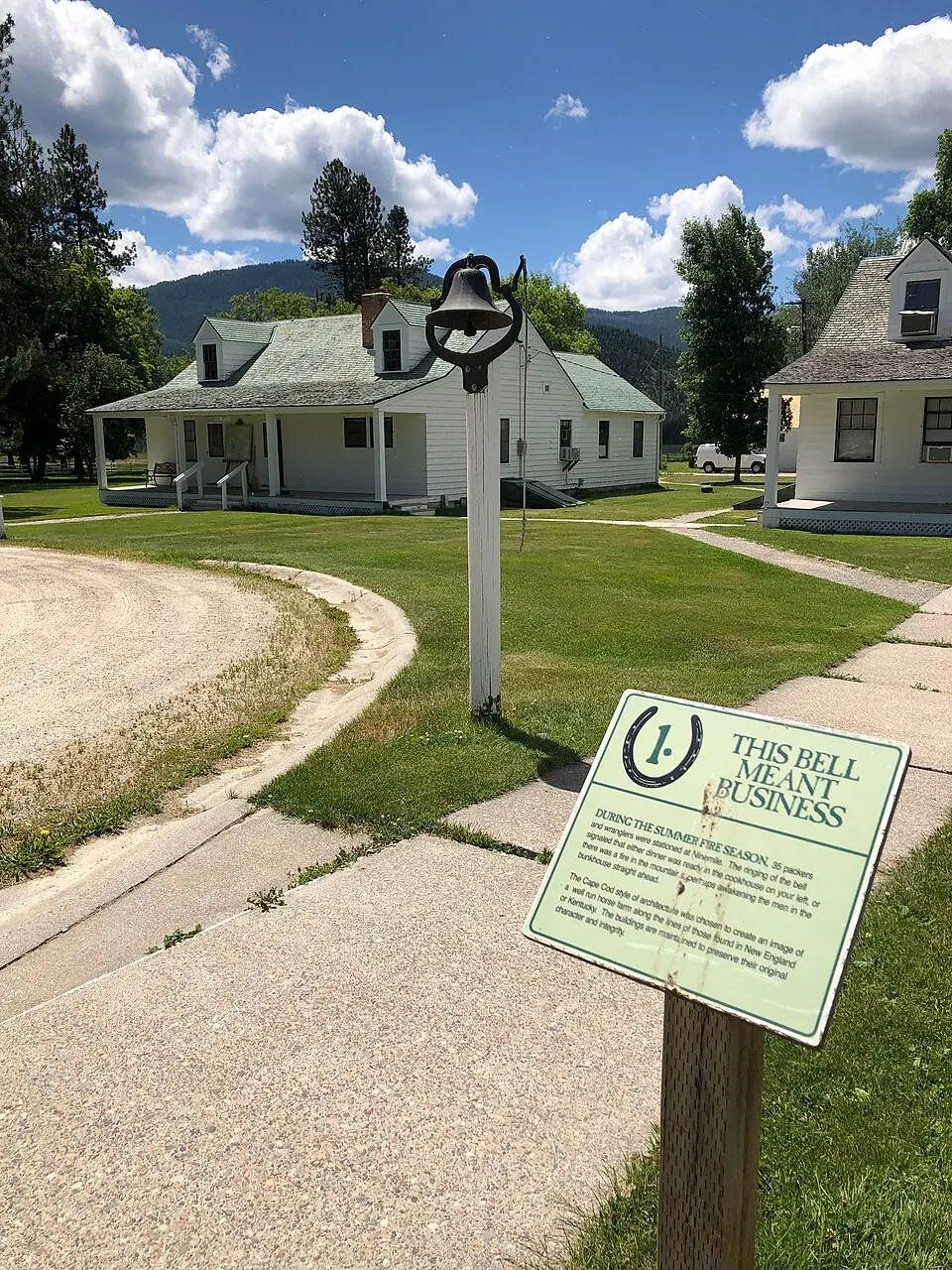 A historical marker stands in front of a white house with a bell mounted on a post.