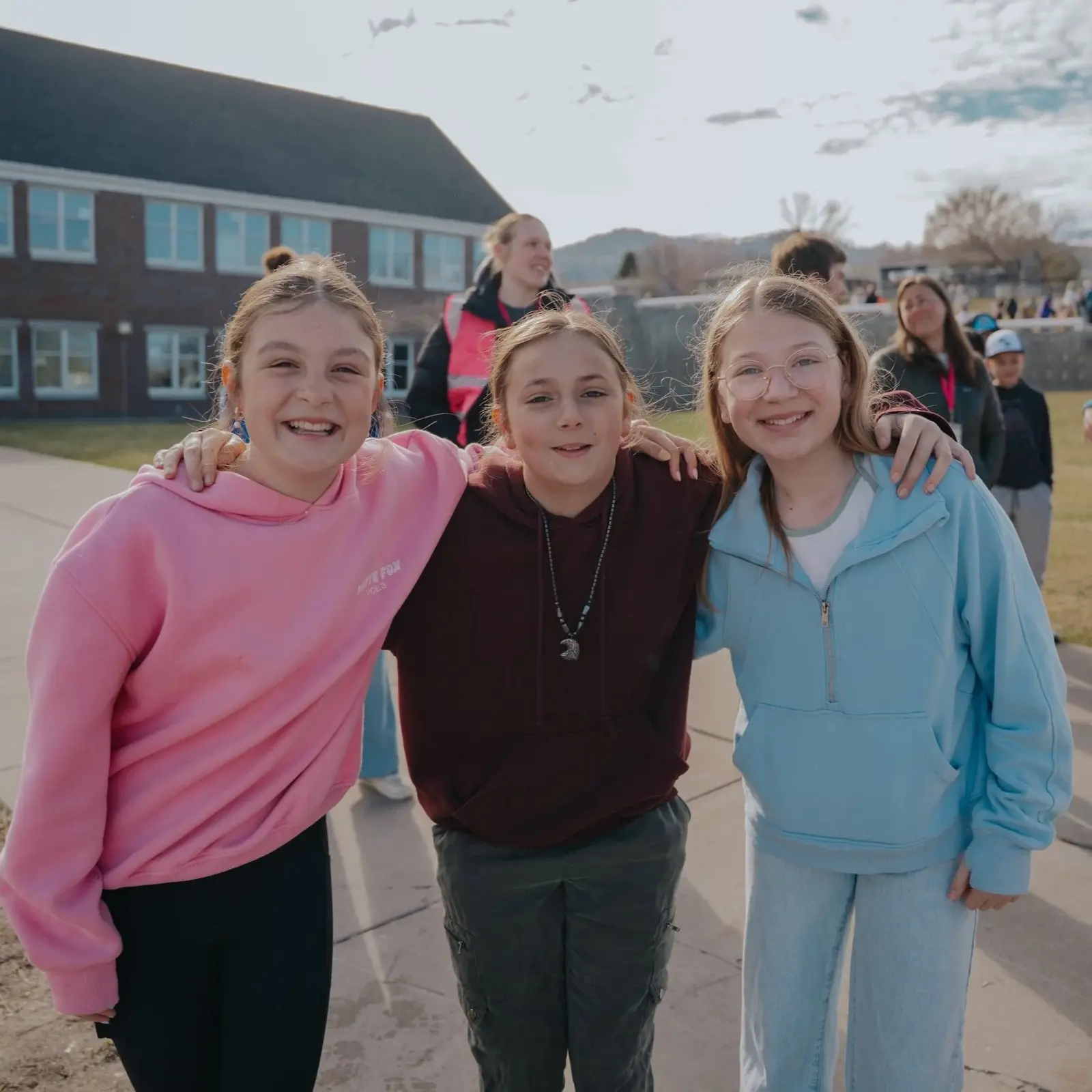 Three girls smiling with their arms around each other outdoors with a school building in the background.