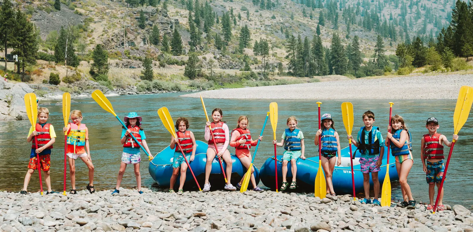 A group of children in life vests stand on a rocky riverbank holding paddles next to two rafts.