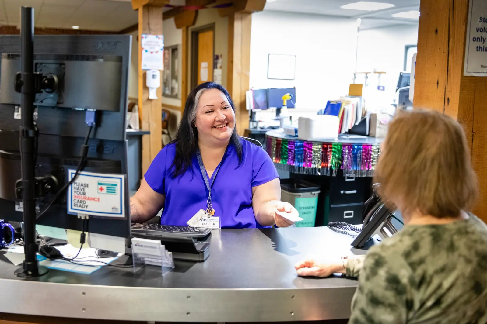 A friendly receptionist at a clinic desk interacts with a patient.