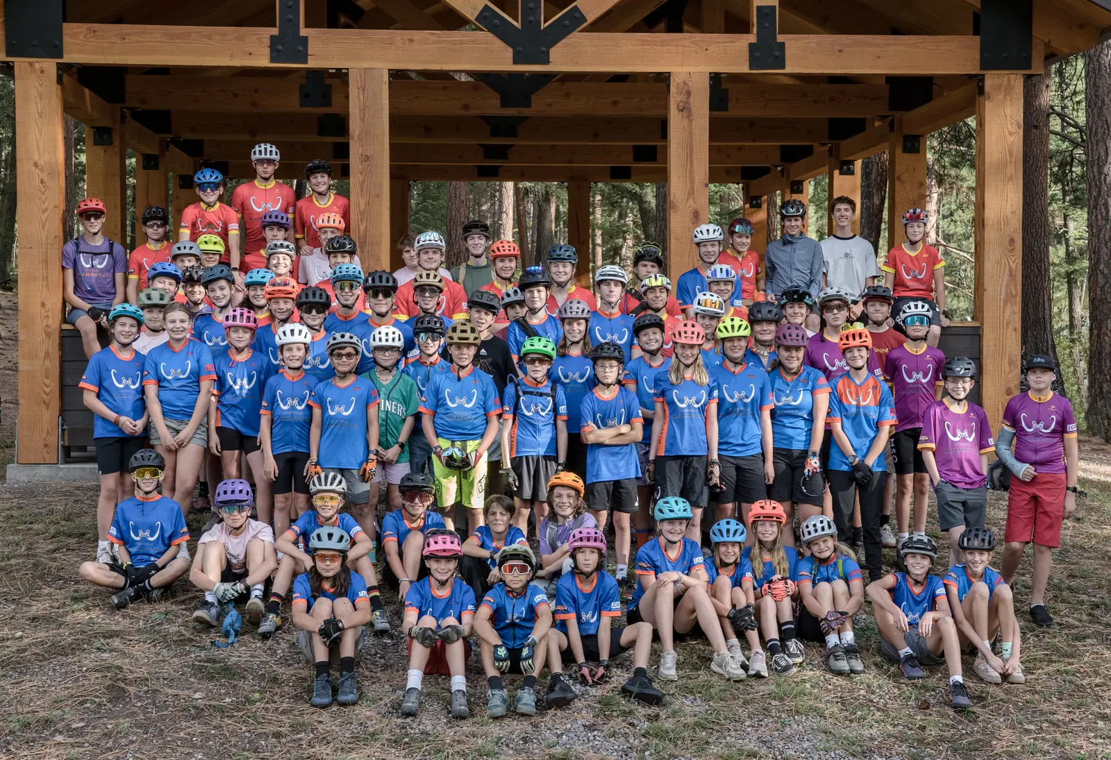 A large group of children and adults pose in cycling gear under a wooden structure.