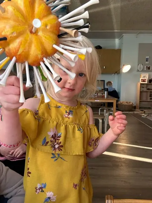 A young girl holds a pumpkin decorated with colorful golf tees in a classroom setting.