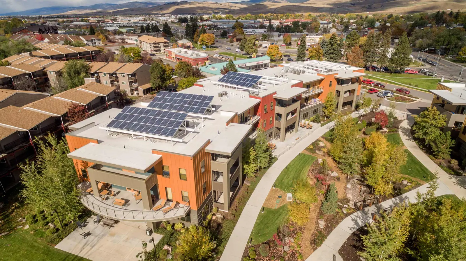 An aerial view shows a modern apartment building with solar panels on the roof surrounded by trees and sidewalks.