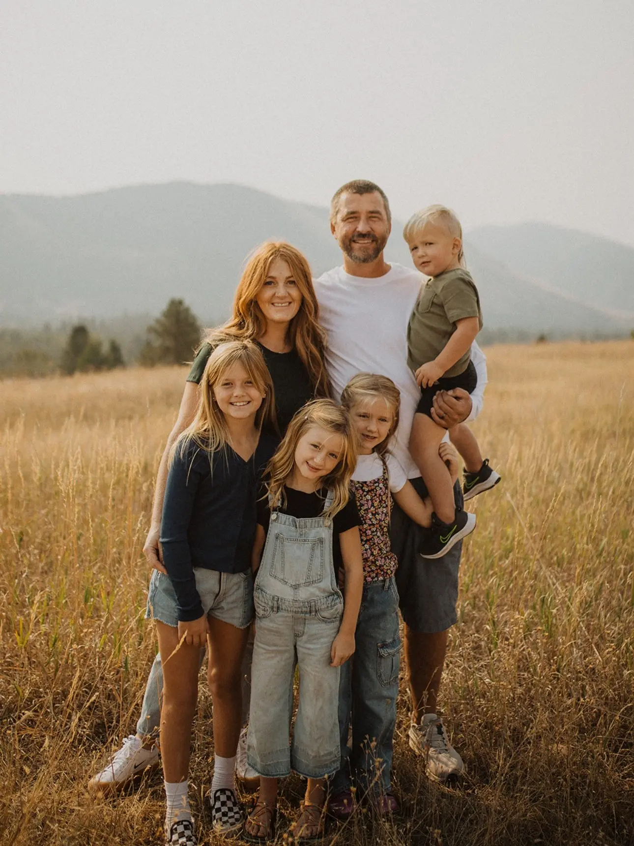 A family of six poses together in a tall grass field with mountains in the background.