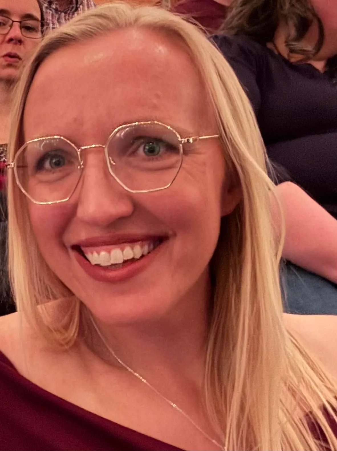 A close-up portrait of a blonde woman wearing glasses and smiling.