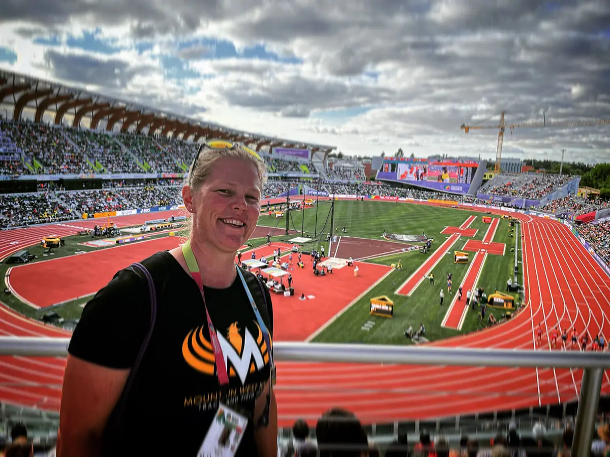 A smiling woman stands in the stadium seating area overlooking a track and field competition.