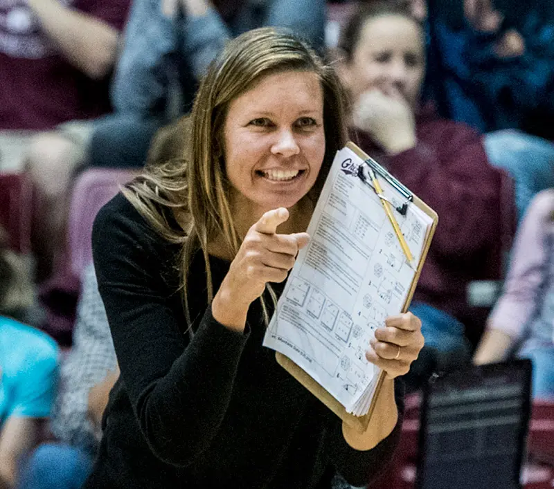 A woman smiles and points while holding a clipboard in front of a crowd.