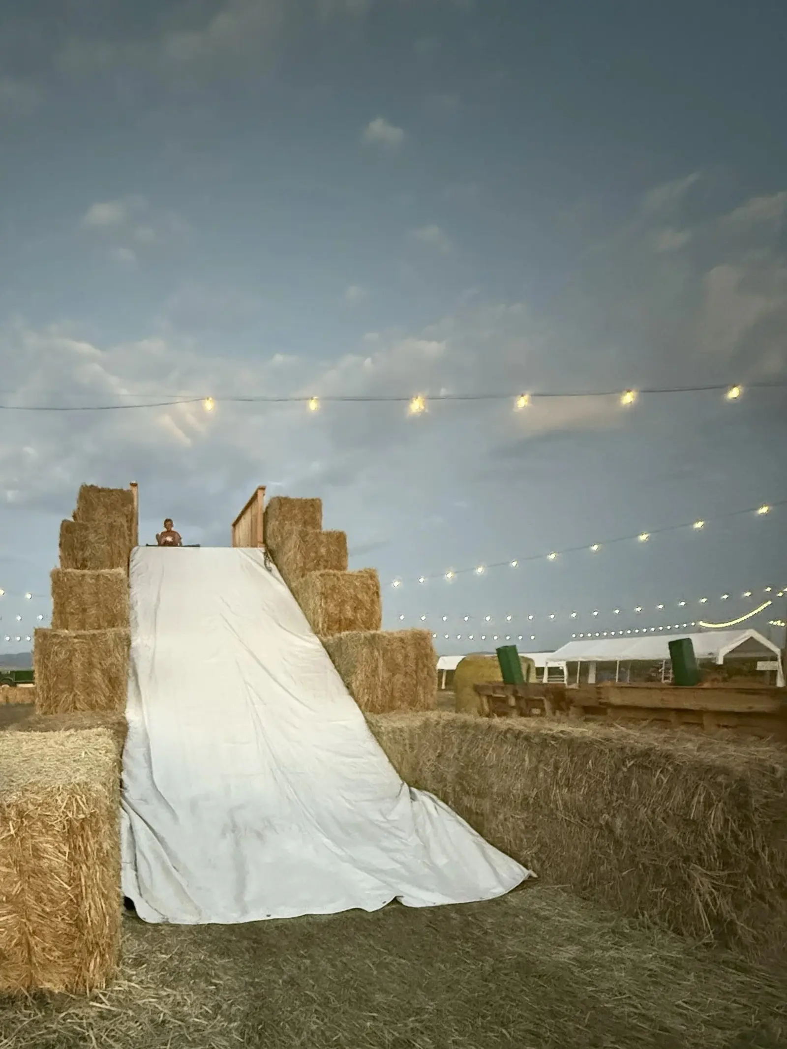 A child prepares to slide down a homemade hay bale slide at an outdoor event under string lights.