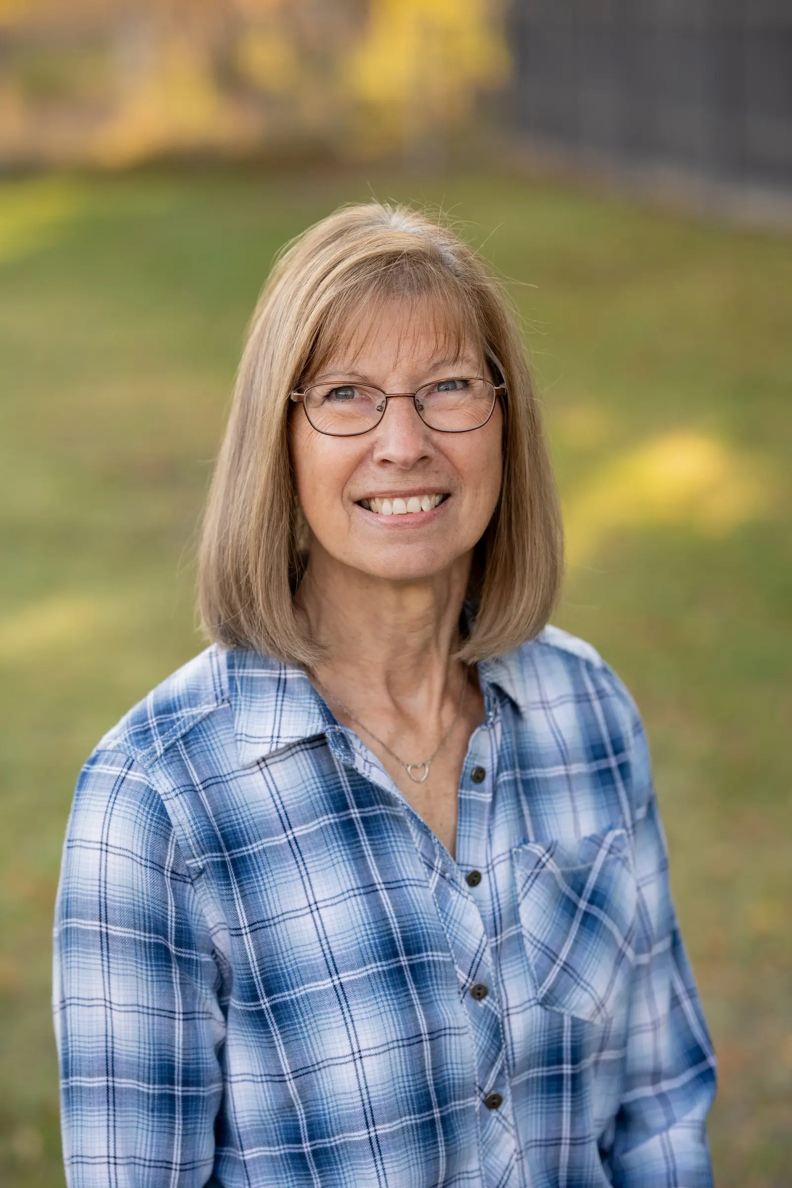 A woman with shoulder-length hair and glasses smiles while wearing a plaid shirt.