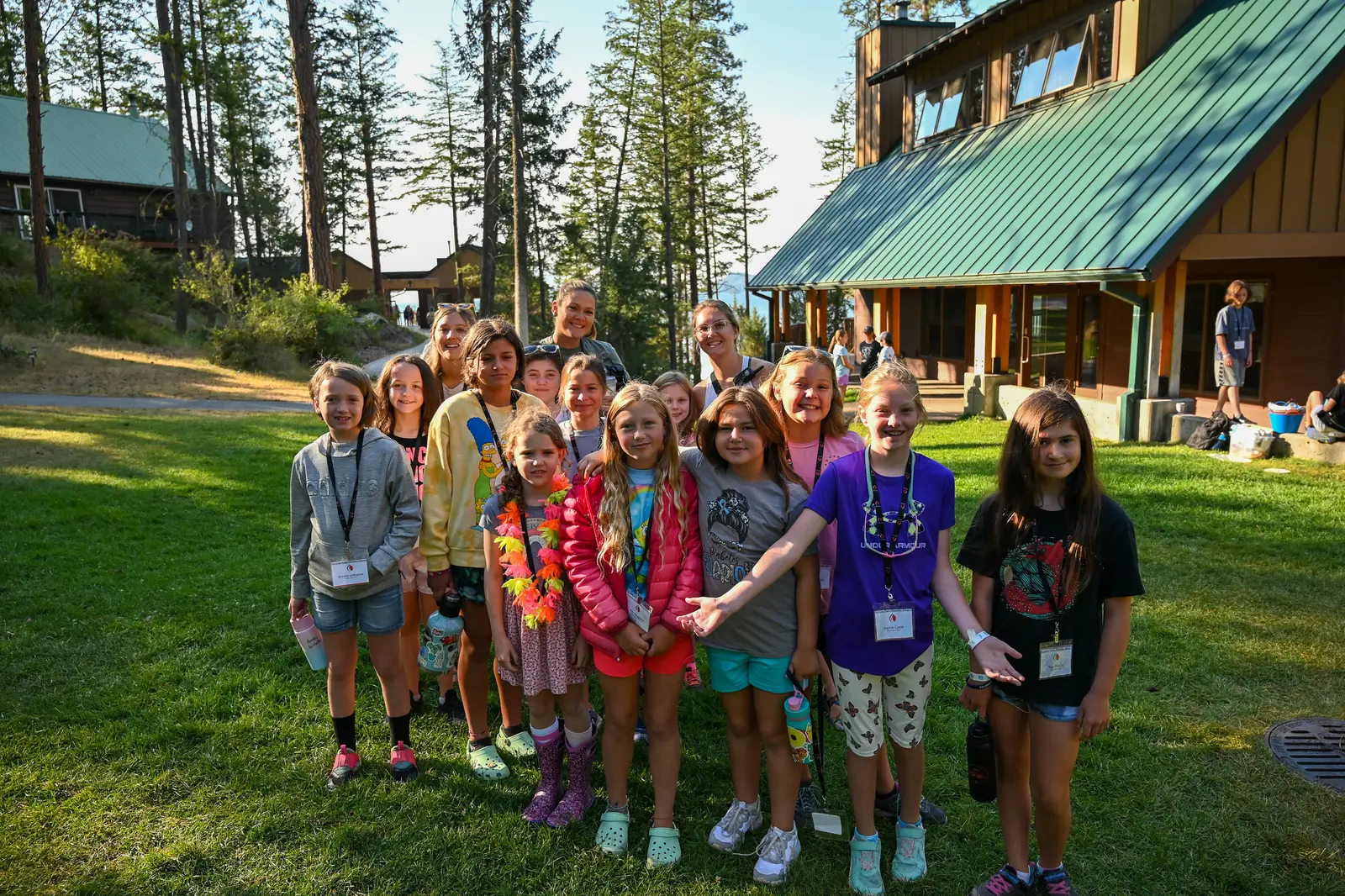 A group of young campers and instructors stand together on a grassy lawn in front of a rustic building.