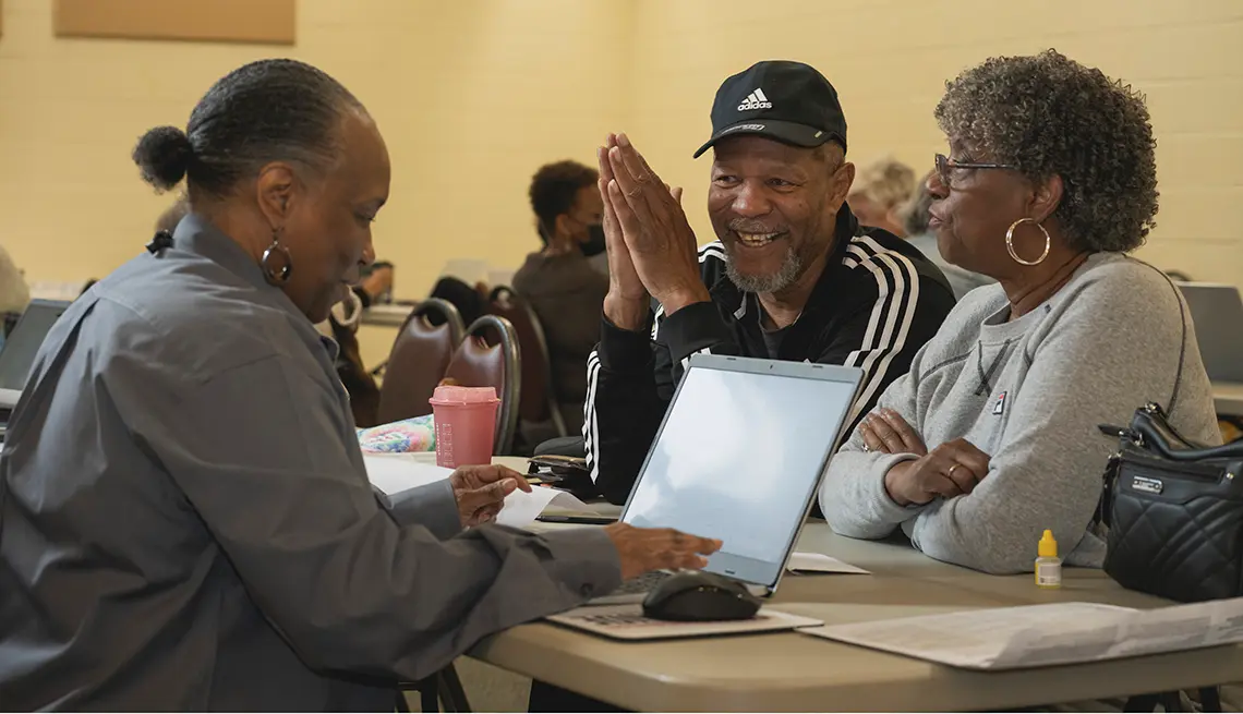 Three adults are gathered around a table with a laptop, with one person clapping.