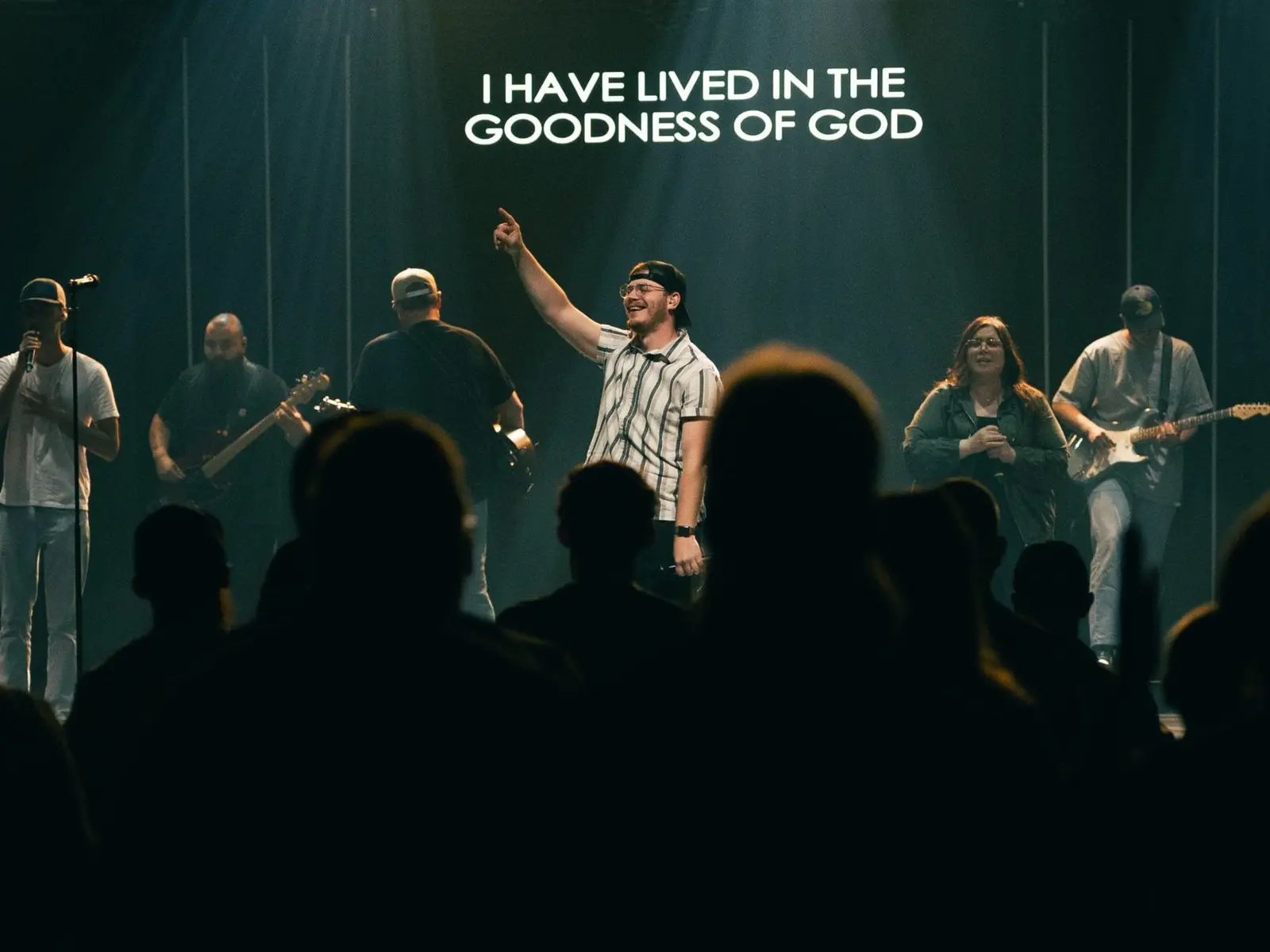 A band performs on a stage in front of an audience with the words "I HAVE LIVED IN THE GOODNESS OF GOD" displayed above them.