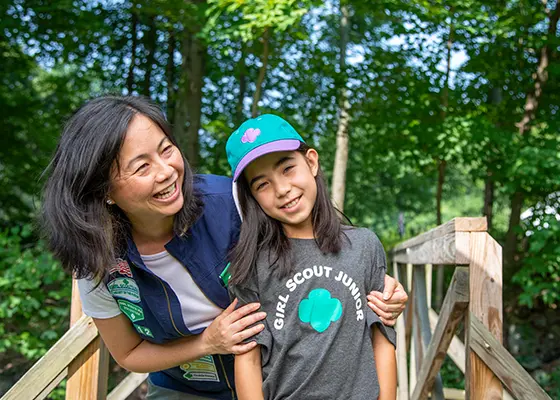 A smiling adult and a child standing together on a wooden bridge in a wooded area.