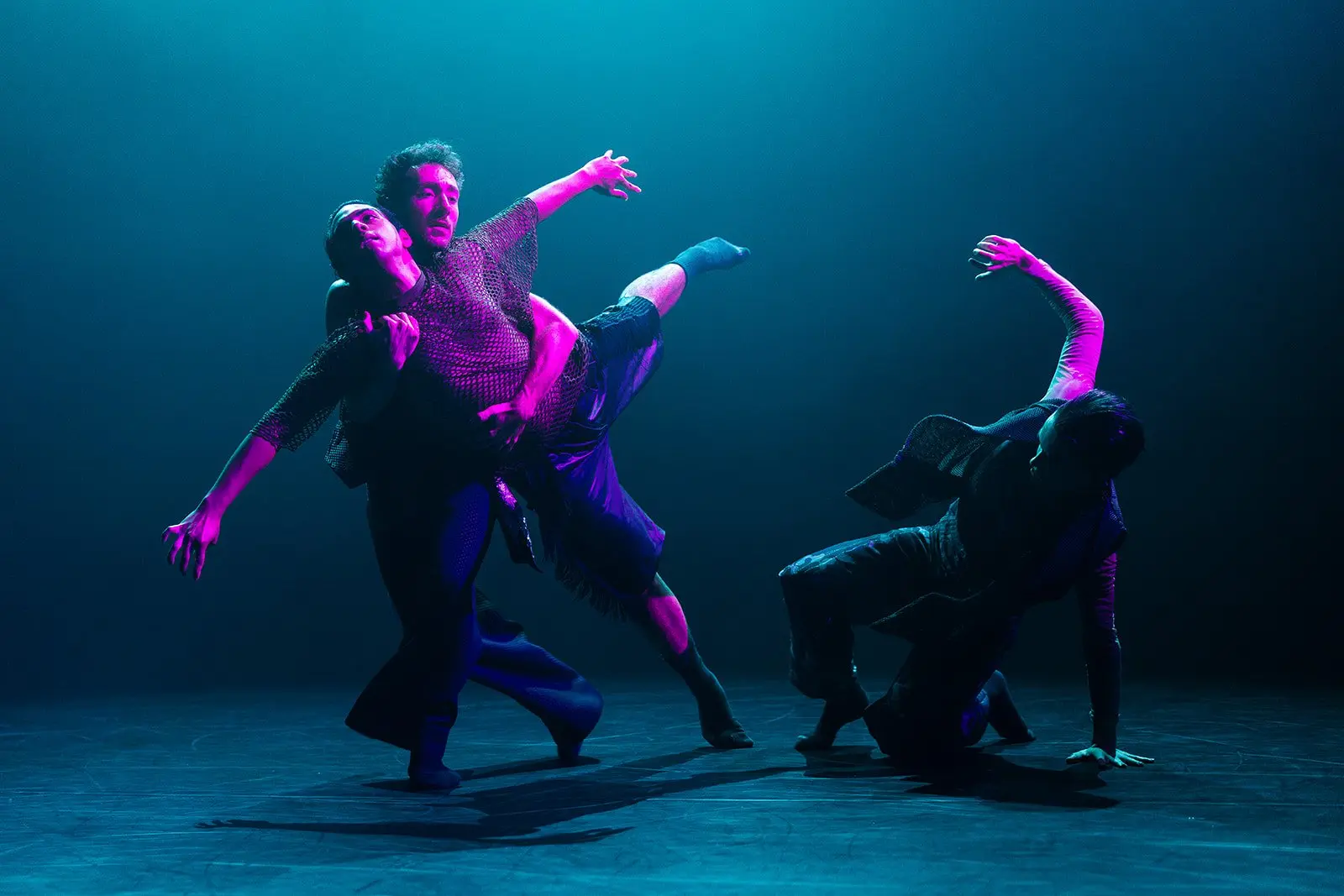 Three dancers perform on a stage under blue and magenta lighting.