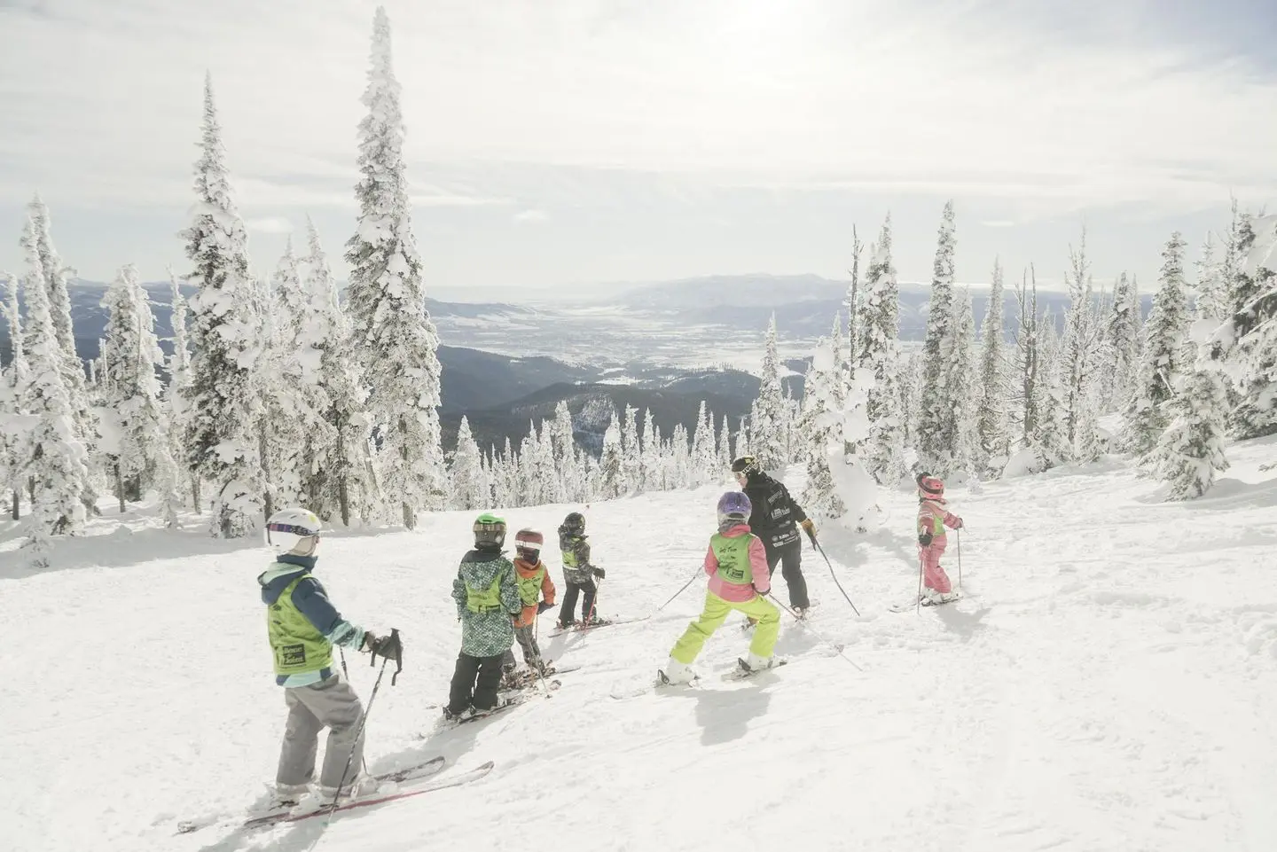 An instructor leads a group of children on skis down a snowy mountain slope.