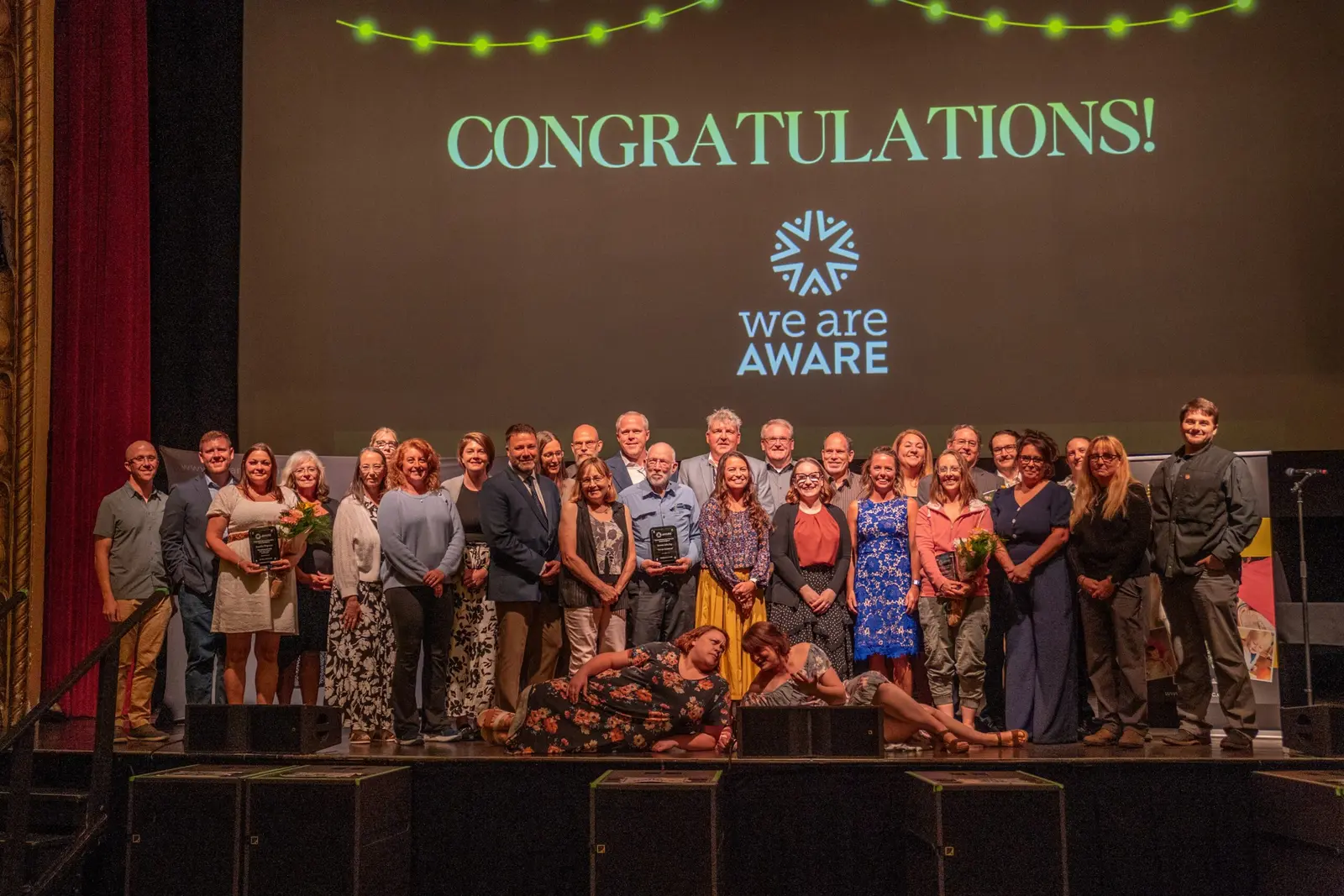 A large group of adults poses for a photograph on a stage under a projection that says CONGRATULATIONS! and WE ARE AWARE.