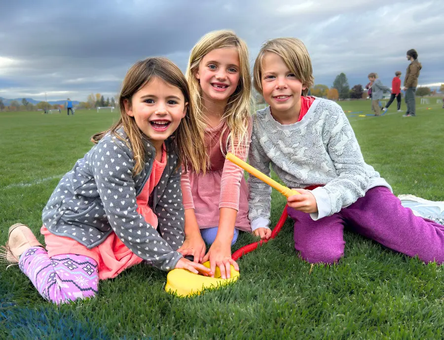 Three children sit on a grass field and play with a yellow toy.