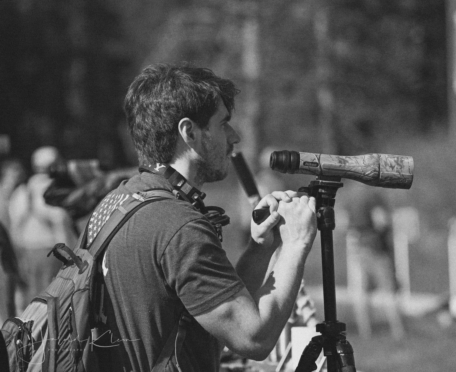 A man uses a spotting scope on a tripod while wearing a backpack.