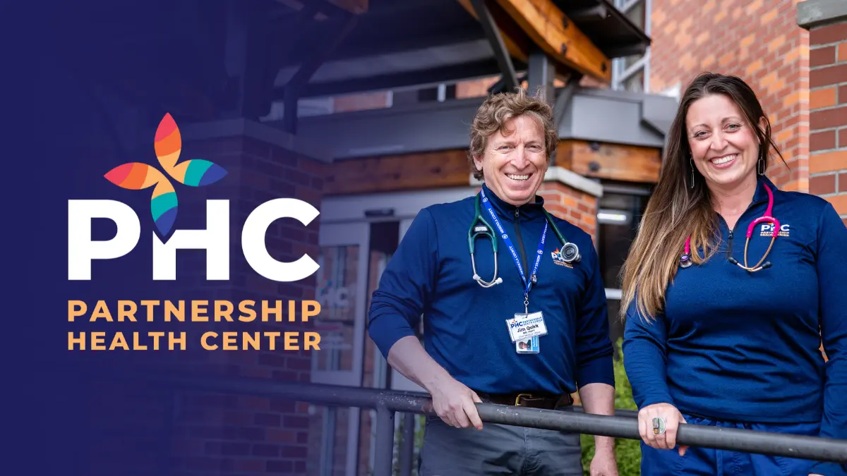 Two medical professionals stand together in front of a brick building with a logo.