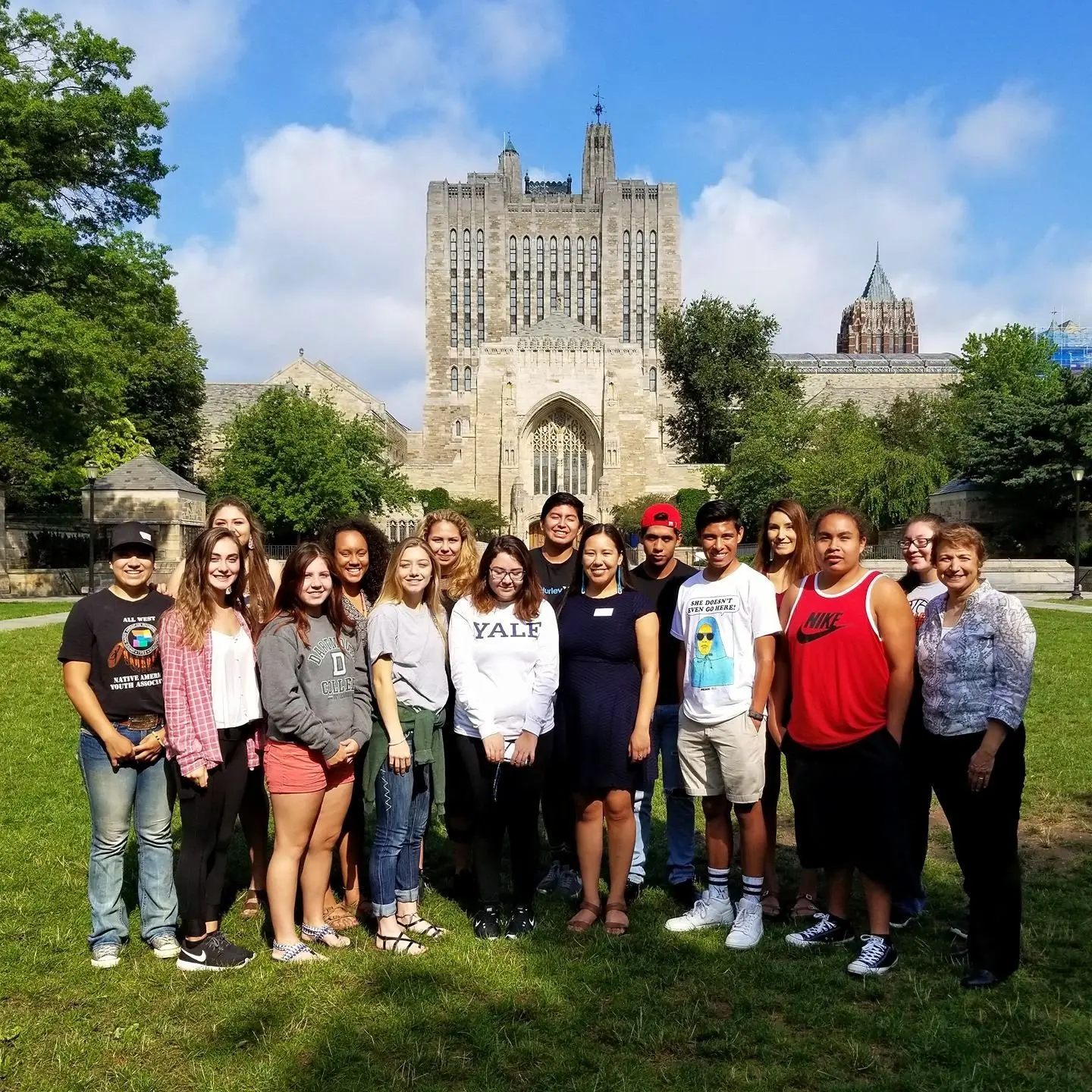 A diverse group of young adults and an instructor stand together on a lawn in front of a large gothic-style stone building.