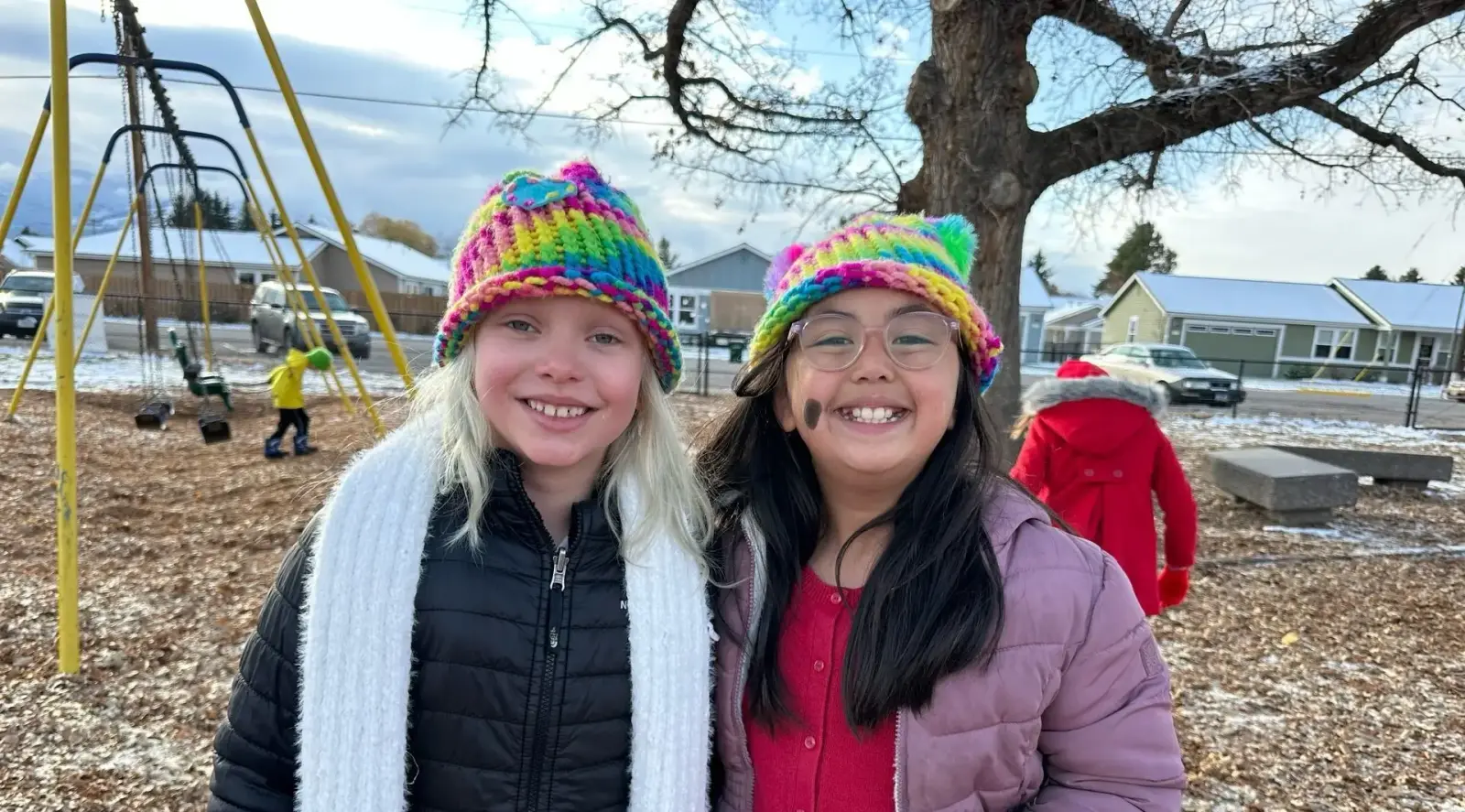 Two children wearing colorful knit hats stand in a park with playground equipment in the background.