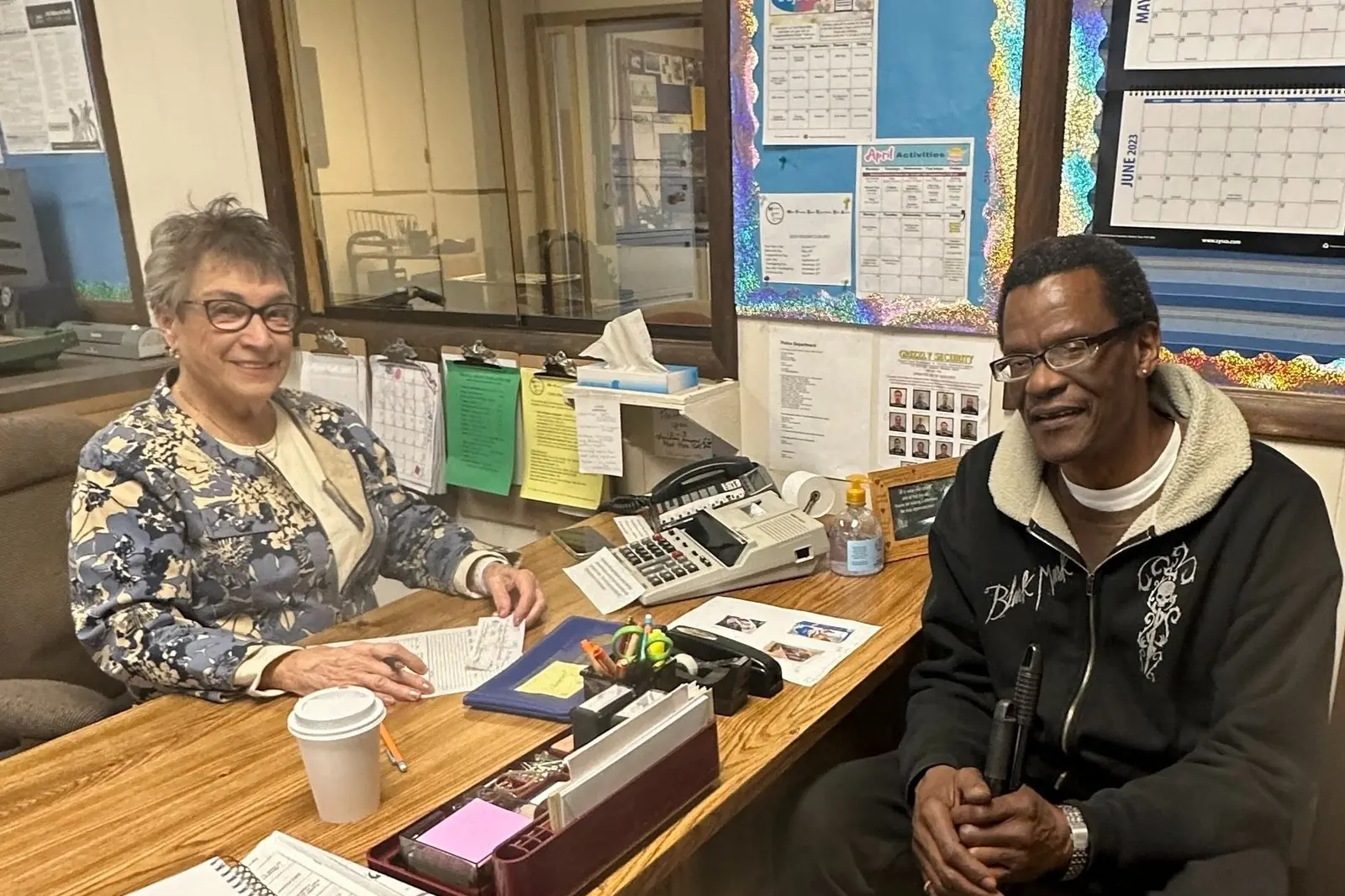 Two people sitting at a desk in an office, smiling and posing for the photo. The woman on the left has short gray hair, glasses, and is wearing a floral jacket. The man on the right has short dark hair, glasses, and is wearing a dark jacket with a beige hood. There are office supplies, papers, a telephone, and a hand sanitizer on the desk. The background has a bulletin board with various papers and a calendar.