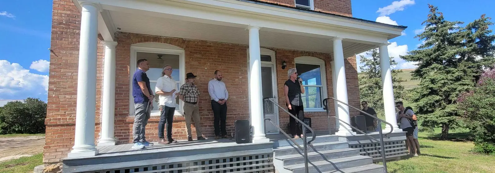 A group of adults stands on the front porch of a brick building as one person speaks into a microphone.