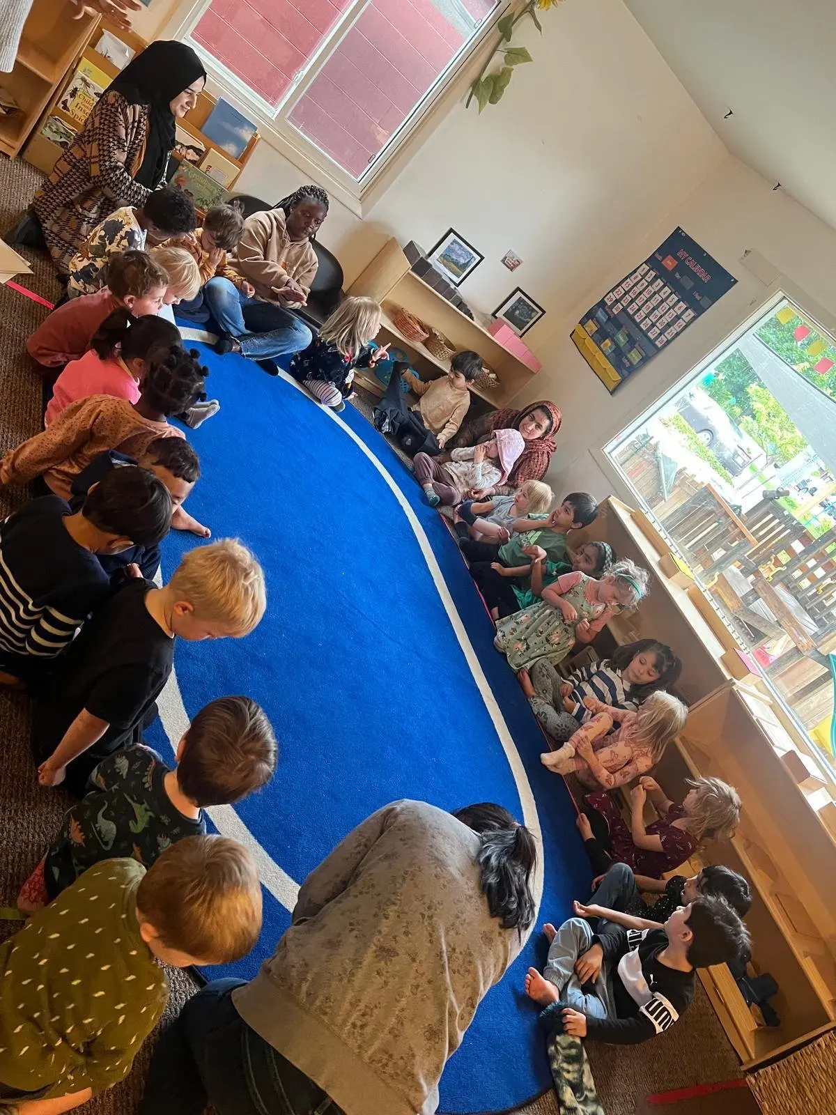 Children and adults sitting and lying around a blue carpet in a classroom.