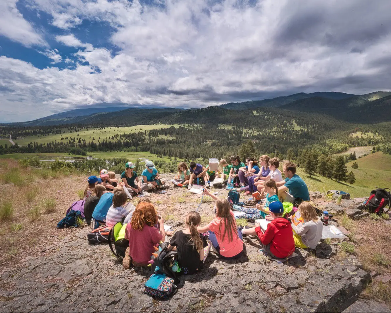 A large group of children and instructors sit in a circle on a rocky hillside overlooking a valley.