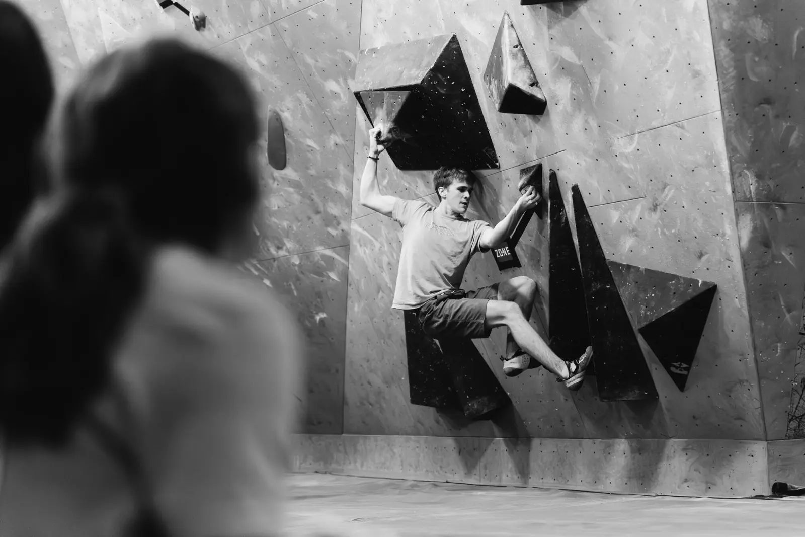 A person rock climbs on an indoor bouldering wall while another person observes from the foreground.