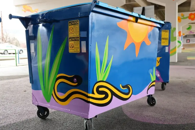 A brightly painted dumpster sits on a paved surface featuring colorful outdoor-themed art.