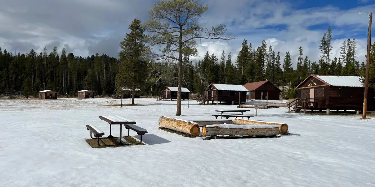 A snow-covered campground features several wooden cabins and outdoor benches.
