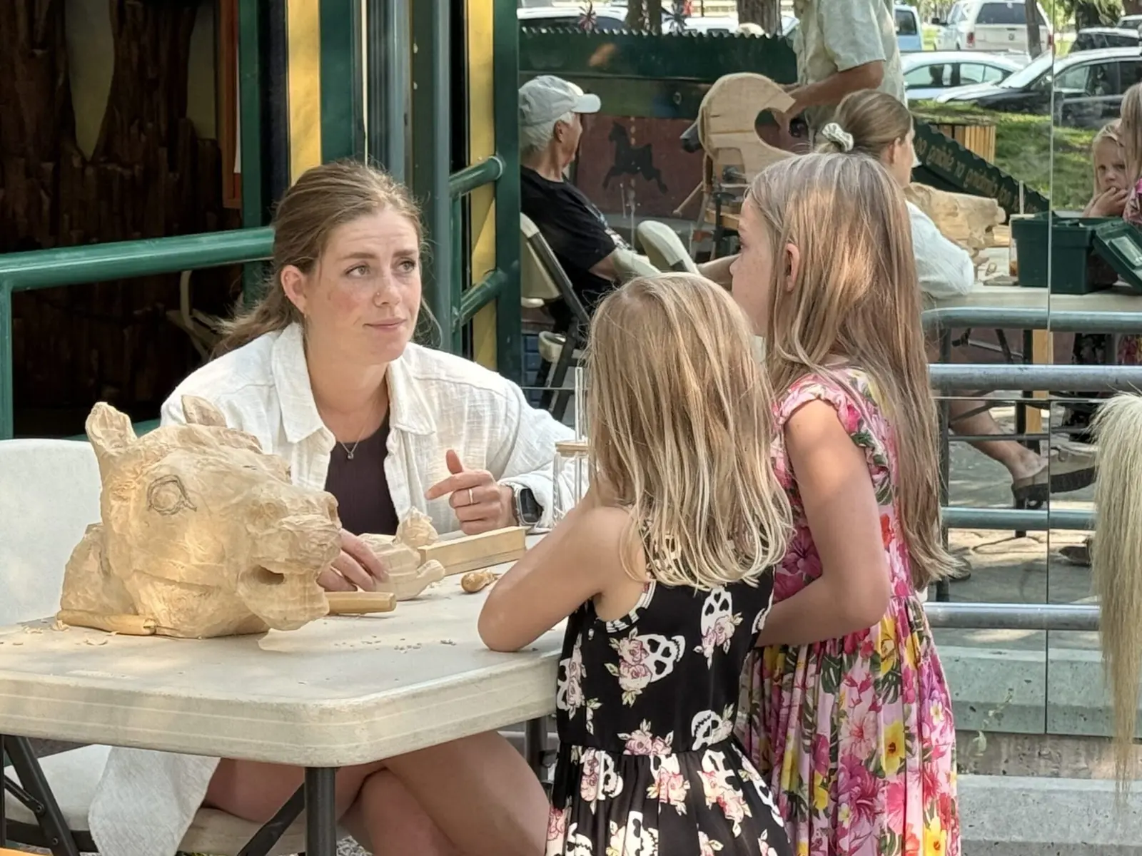 A woman demonstrates a wood-carving project to two young girls at a table.