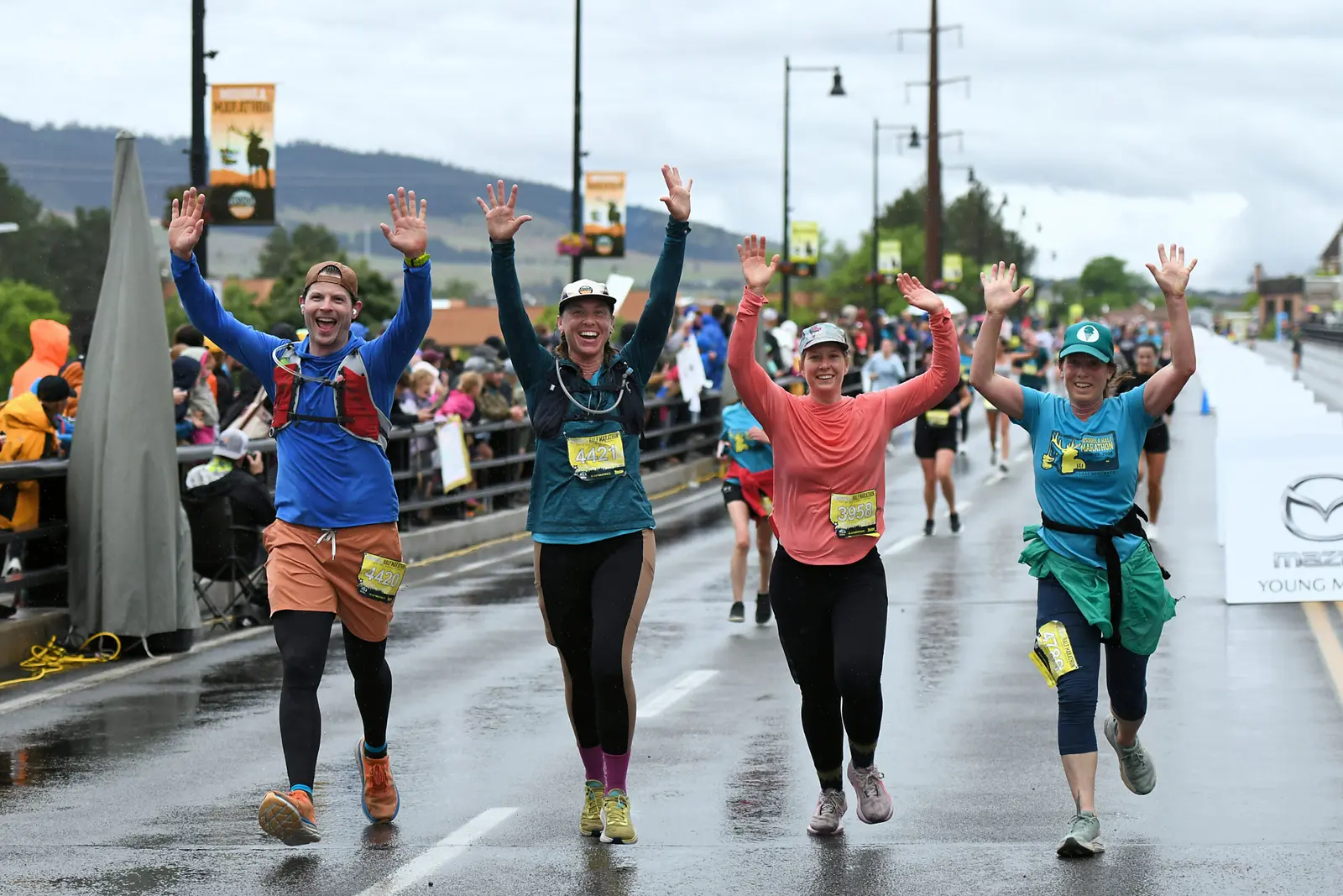 Four runners with arms raised celebrate while running on a wet city street during a marathon.
