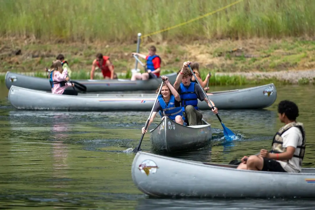 A group of young people paddle canoes on a lake.