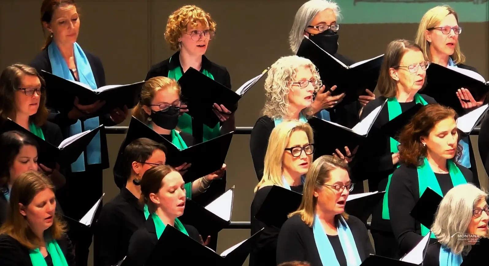 A choir of women singing, wearing black attire with teal and green scarves, some wearing glasses, some wearing face masks, holding sheet music, singing in a concert hall.