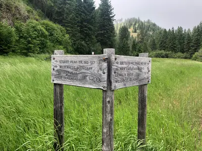 Photo from fs.usda.gov A wooden trail signpost stands in a grassy field with forested hills in the background.