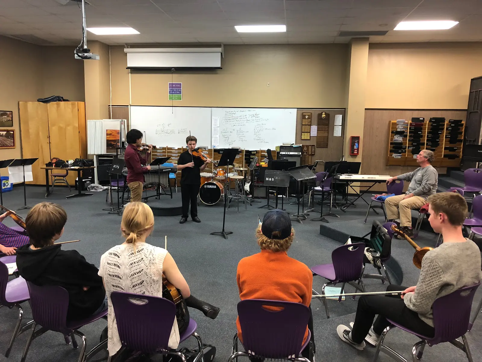 A student plays the violin in a classroom while other students and an instructor watch.
