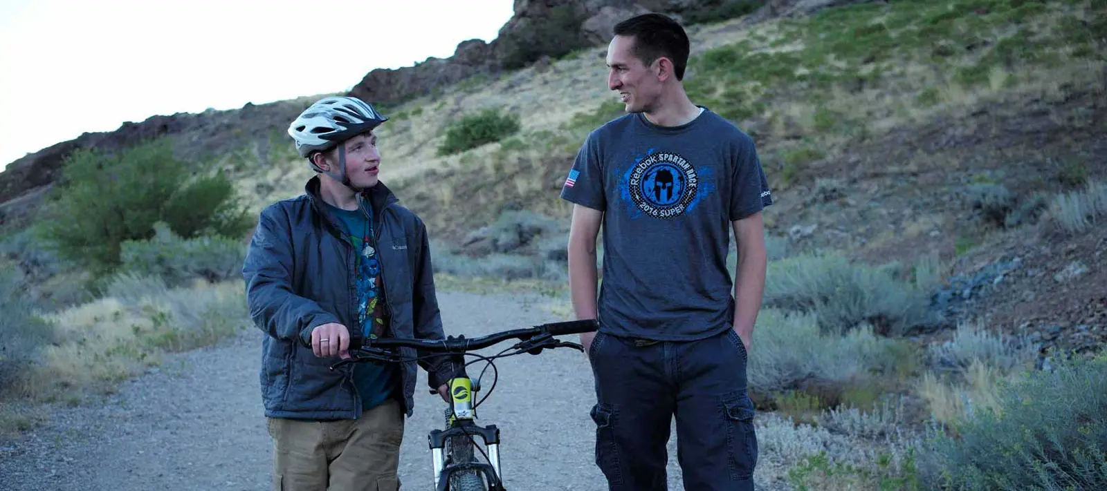 Two young men walk together on a dirt trail, one with a bicycle.