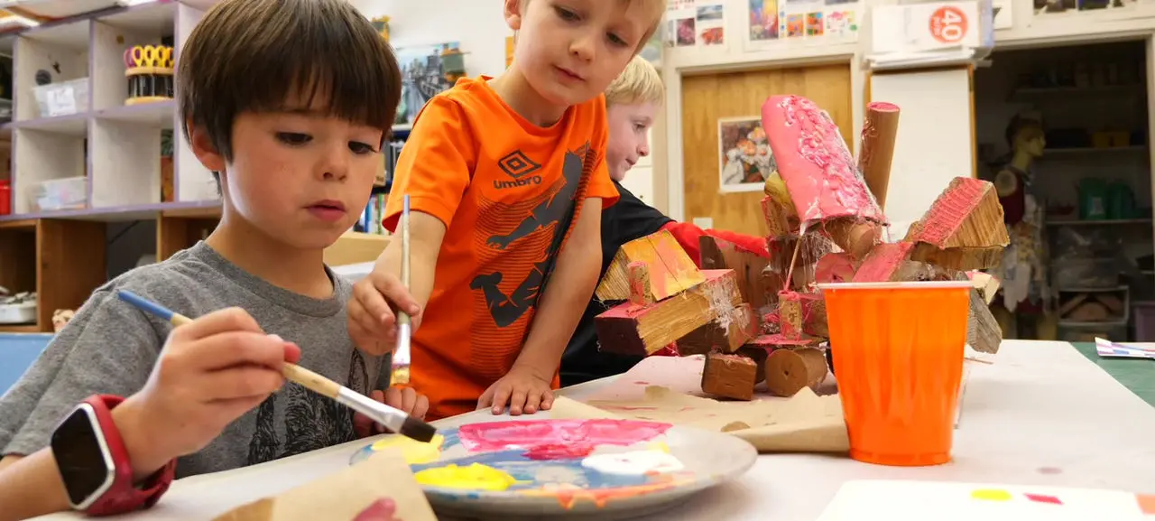 Two young boys are focused on painting an abstract wooden sculpture with colorful paint.