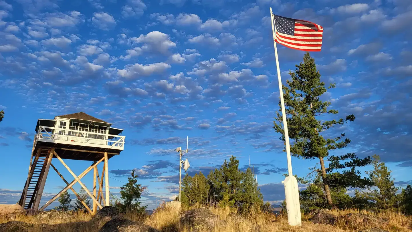 Double Arrow Lookout Tower- Tower in background, flag pole with American flag in foreground, fluffy grey clouds pass over a partly cloudy blue skyDouble Arrow Lookout- Facing West